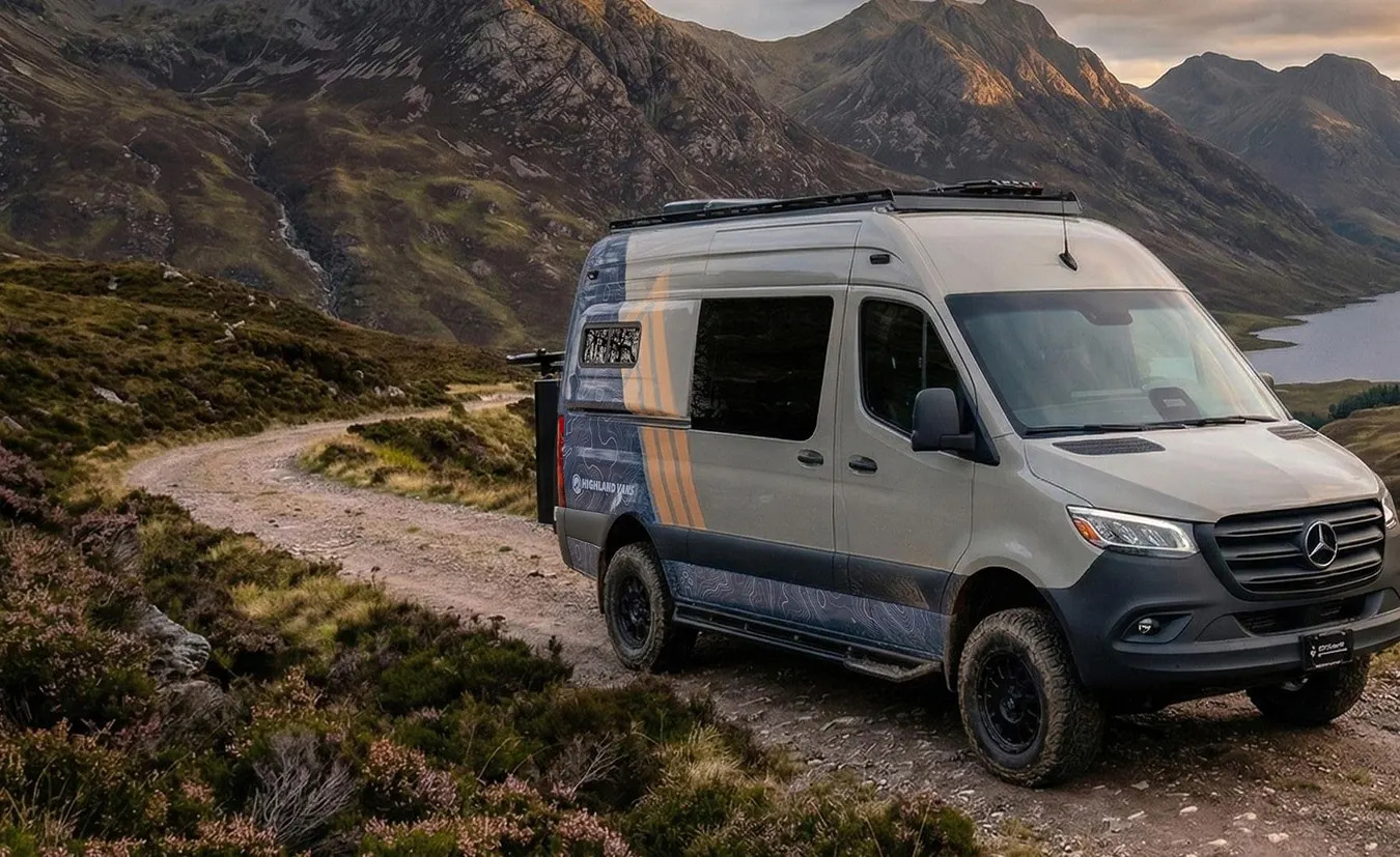 A Highland Vans Sprinter in sand and blue topographic livery, parked on a mountain track above a loch at sunset
